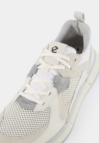 Leichter Sneaker mit einem Obermaterial aus Mesh und Wildleder in Weiß und Grau, ausgestattet mit einem gepolsterten Knöchelbereich, einer Schnürung und strukturierten Akzenten.