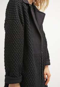 Abrigo negro con una tela texturizada, similar a burbujas, y un cuello y puños lisos de contraste. Manga larga y diseño de apertura frontal.