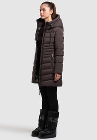 Brown puffer coat featuring a hood, quilted design, front zipper, side pockets, and ribbed cuffs; paired with glossy black snow boots.