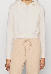 Donna che indossa una felpa crema con zip e un anello per la zip, insieme a pantaloni beige con coulisse a costine, in piedi con le mani rilassate ai lati.