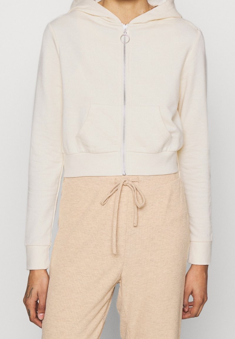 Donna che indossa una felpa crema con zip e un anello per la zip, insieme a pantaloni beige con coulisse a costine, in piedi con le mani rilassate ai lati.