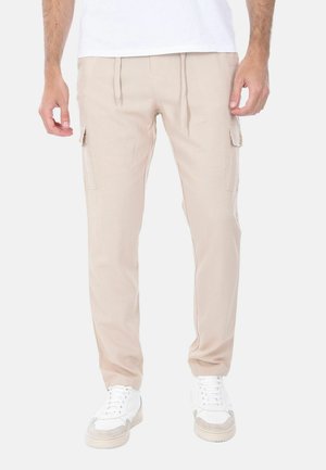 Uomo che indossa pantaloni cargo beige e scarpe da ginnastica bianche, in piedi con le mani rilassate lungo i fianchi su uno sfondo bianco uniforme.