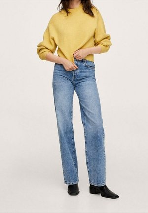 Frau trägt einen gelben Strickpullover, hoch geschnittene gerade blaue Jeans und schwarze Stiefeletten und justiert den Saum des Pullovers.