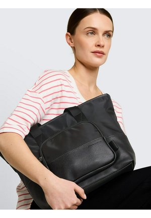 Femme portant une chemise blanche à rayures rouges, tenant un grand sac cabas noir avec une poche zippée à l'avant sur un fond uni.