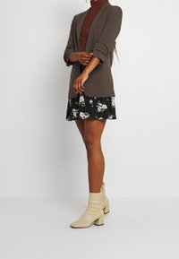 Brun blazer över en vinröd polotröja, kombinerad med en svart blommig kjol och kremfärgade ankelboots med klack. Släta tygtexturer är tydliga.