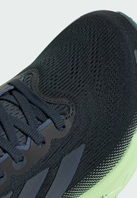 Černé sportovní boty s texturovaným pleteným svrškem, černými tkaničkami, šedými akcenty a světle zelenou podrážkou se proužkovanými vzory.