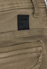 Pantalones caqui con una etiqueta de cuero negro que lleva el texto "ALPHA INDUSTRIES", costuras cosidas y un acabado de tela texturizada.
