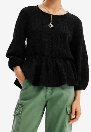 Kvinde iført tekstureret sort peplum-top med lange ærmer, grønne cargobukser og en guldkæde med en sommerfuglevedhæng.