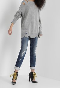 Pull surdimensionné gris avec détails usés, associé à un jean bleu déchiré et des bottes à talons noires avec des lacets jaunes.