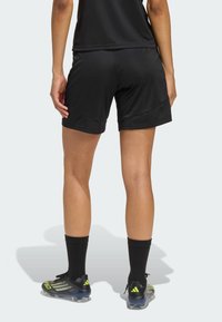 Μαύρο αθλητικό σορτς με λείο υφή, ελαστική μέση και πλευρικές ραφές· φορεμένο με μαύρες κάλτσες και αθλητικά παπούτσια με πράσινες λεπτομέρειες.