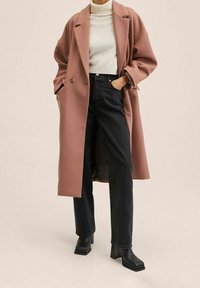 Une femme porte un manteau long rose poudré, un pull à col roulé blanc, un jean noir taille haute et des bottines à talons carrés noires, debout les mains dans les poches.