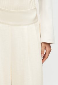 Το χέρι μιας γυναίκας στο πλάι, φοράει λευκή μπλούζα με μακριά μανίκια και ψηλόμεσο, φαρδύ, κρεμ παντελόνι με λεία μέση.