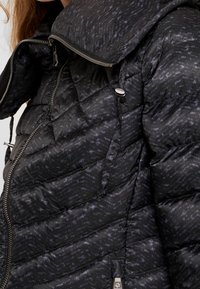 Kvadricerat, svart dunjacka med hög krage, silvriga dragkedjor och ett subtilt mönster; glänsande tyg med en mjuk textur.