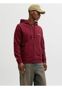 Sweat à capuche bordeaux zippé avec une poche avant, une capuche à cordon de serrage et un logo brodé en or. Porté avec un jean gris clair.