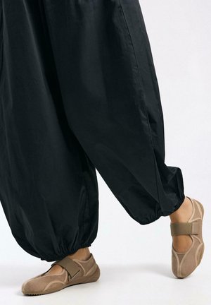 Fekete, szűkülő szabású nadrág, rugalmas szegélyekkel, bézs színű vászon cipőkkel párosítva, amelyek tépőzáras pánttal és texturált oldalsó díszítéssel rendelkeznek.