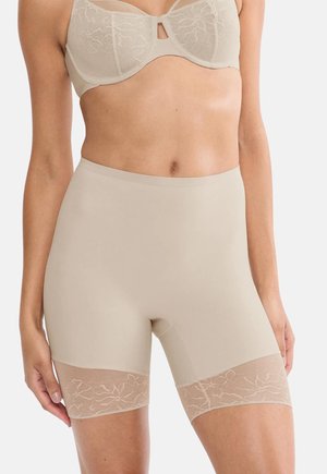 Žena oblečená v béžových tvarujúcich krátkych nohavičkách s vysokým pásom a čipkovaným lemom a zodpovedajúcej čipkovanej podprsenke s kostícami, stojaca pred jednoduchým pozadím.