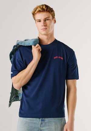 Navyblå bomuld T-shirt med korte ærmer, med rød "PEPE JEANS" logo på brystet. Båret med en denimjakke hængende over skulderen.