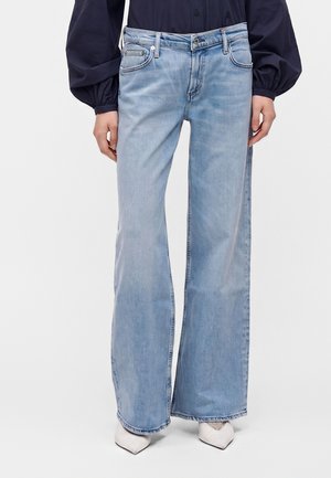 Personne portant un jean bleu clair à jambes larges avec un chemisier à manches longues foncé et des chaussures blanches à bout pointu, debout devant un fond uni.