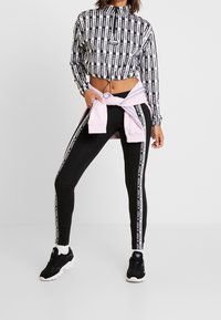 Donna che indossa leggings neri Adidas con strisce con logo, un top cropped bianco e nero Adidas, scarpe da ginnastica nere e una felpa rosa annodata.