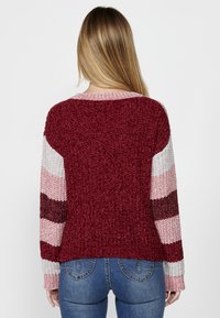 Pleteni pulover z maroon telesom in kontrastnimi črtastimi rokavi v roza, burgundy in sivi barvi. Teksturovana tkanina z ribanim robom in manšetami.