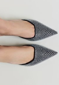 Ballerines noires à bout pointu ornées de strass argentés, dessus texturé et bordure noire contrastante, reposant sur un fond neutre.