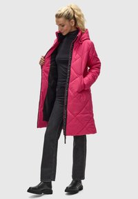 Lunga giacca in piuma rosa trapuntata con cappuccio, dotata di cerniera nera e fodera interna. Abbinata a un top nero e jeans grigi. Stivali neri alla caviglia.