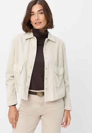 Frau trägt eine beige Cordjacke über einem dunkelbraunen Rollkragenpullover und beigen Hosen mit braunem Gürtel, steht vor einem einfachen weißen Hintergrund.