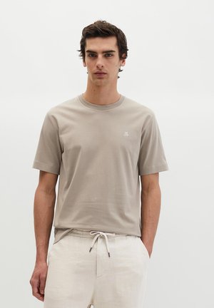 Jonge man staat met zijn handen in zijn zakken, draagt een beige T-shirt met korte mouwen en lichte crèmekleurige broek met trekkoord tegen een effen achtergrond.