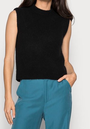 Kobieta ubrana w bezrękawnik czarny, puszysty sweter, zestawiony z wysokim stanem turkusowych spodni, jedna ręka w kieszeni, kadrowana w okolicach brody i ud.