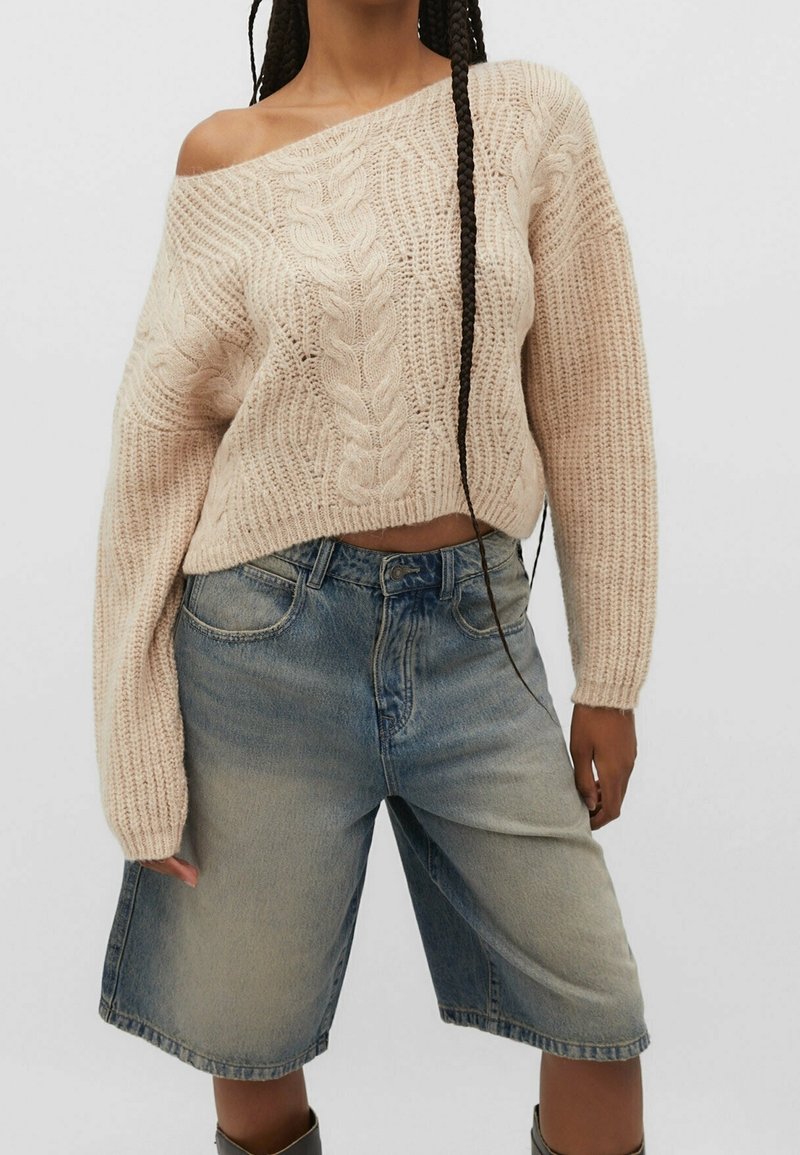 Maglione lavorato a maglia color crema con motivo a treccia e maniche ampie, abbinato a pantaloni corti in denim chiaro. Il maglione è di stile off-shoulder.