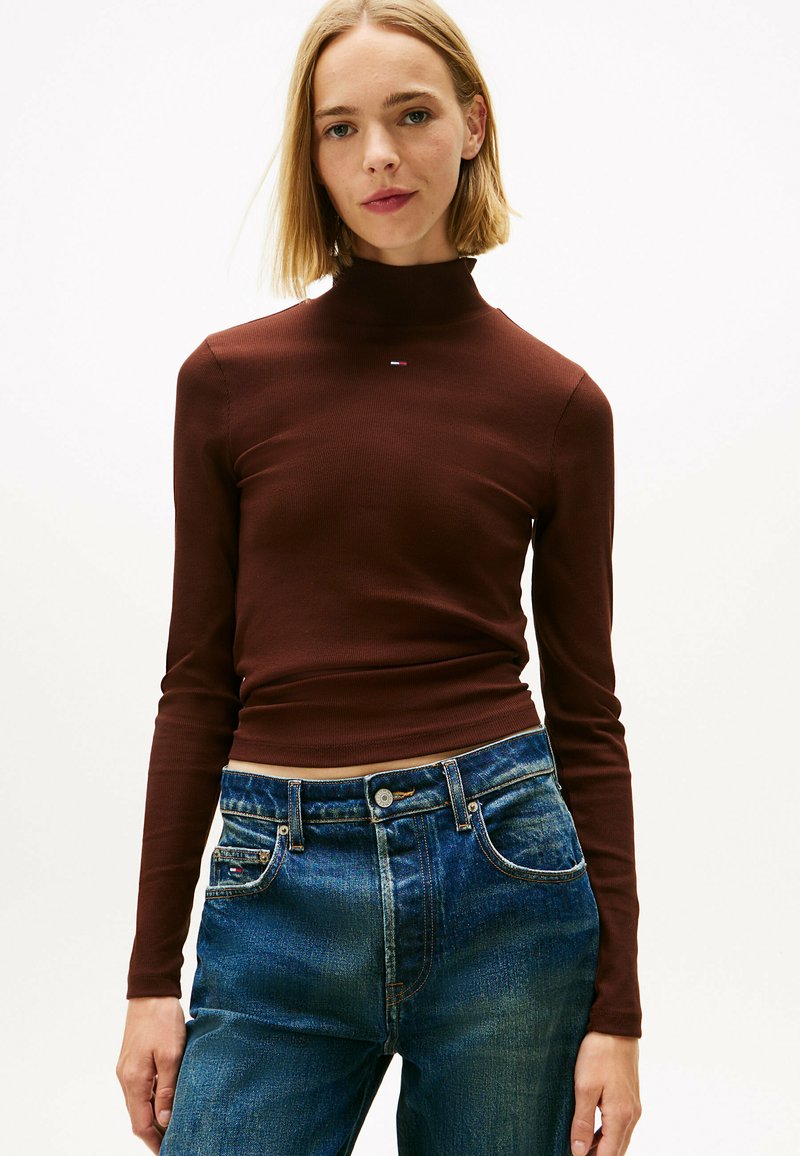 Maglione lungo marrone a maniche lunghe in materiale a coste con colletto alto, abbinato a jeans in denim blu con tasche classiche e chiusura a bottone.