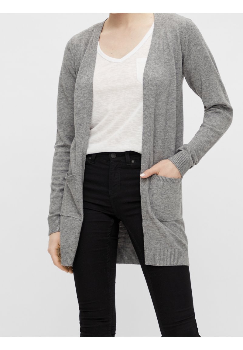 Woman wearing a light grey open-front cardigan with front pockets over a white T-shirt and black pants, hand in cardigan pocket.