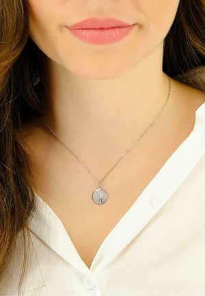 Collier en argent avec un pendentif rond, arborant un design gravé, sur une chaîne délicate. Porté sur une chemise blanche à col.