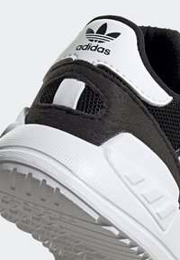 Svart och vit idrottssko med ett övre i nätmaterial, logotyp på hälen och en texturerad vit gummi sula med framträdande mönster för grepp.