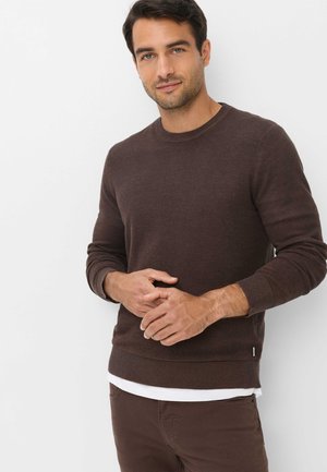Mand iført en brun strikket sweater over en hvid skjorte og brune bukser, stående med hænderne foldet foran en hvid baggrund.
