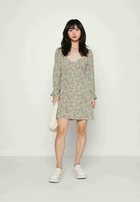 Vestido floral verde com mangas bufantes longas, decote quadrado e um corte solto, combinado com sapatos plataformas brancos e uma bolsa texturizada.