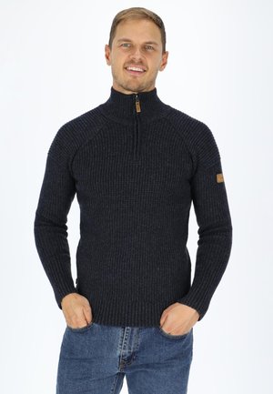 Dark navy ribbed knit sweater with a high collar, half zip closure, and elbow patches, paired with blue jeans. Textured fabric detail visible.