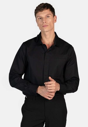 Black long-sleeve shirt made of smooth fabric, featuring a collared neck, front pocket, and fitted design. Worn with black trousers.
