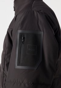 Czarna pikowana rękaw kurtki z kwadratową kieszenią na suwak, ozdobioną wytłoczonym logo BOSS na górnej części ramienia.