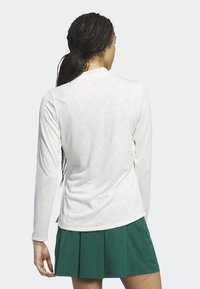 Bílý top s dlouhým rukávem s texturovaným vzorem, kulatým výstřihem a bočními rozparky, doplněný zelenou plisovanou sukní. Hladká textilie.