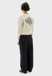 Personne avec les cheveux courts portant un pull crème avec des motifs de fleurs noires dans le dos et un pantalon cargo noir ample, se tenant devant un fond uni.