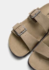 Bruna mocka sandaler med två justerbara remmar, med silverfärgade metallspännen och en texturerad fotbädd. Platt sula med en lätt kurva.