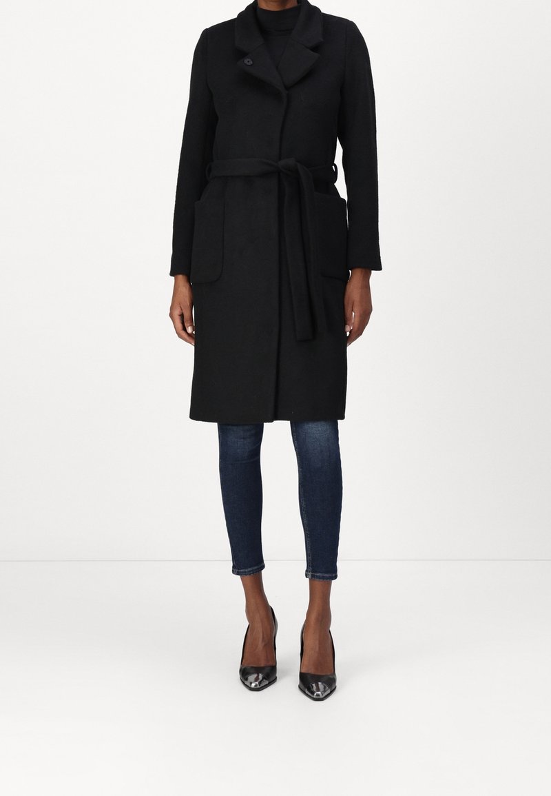 Femme portant un manteau sans manches noir ceinturé par-dessus un top à manches longues, un jean skinny foncé et des escarpins noirs à bout pointu, debout sur fond blanc.