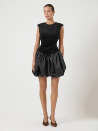 Mulher em pé, vestindo um vestido preto sem mangas com um corpete justo e uma saia volumosa texturizada, combinando com sapatos de salto agudo castanhos.