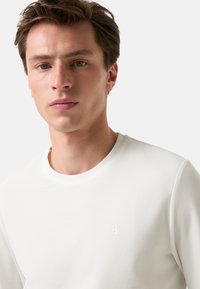 Biała koszulka typu crew-neck z lekkiego materiału. Posiada subtelne wytłoczone logo "B" na klatce piersiowej. Gładka faktura, dopasowany krój i minimalne detale.