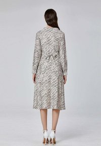 Robe légère en crème avec un motif quadrillé noir en vague, manches longues et design à taille nouée, associée à des sandales à talons hauts blanches.