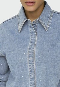 Light blue denim shirt, featuring decorative silver studs, a pointed collar, and front button closure. Textured fabric with visible stitching.