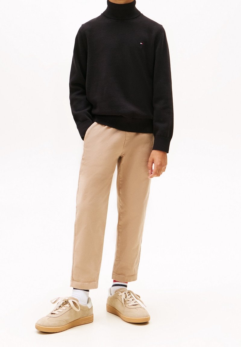 Personne porte un pull à col roulé noir, un pantalon beige raccourci, des chaussettes blanches et des baskets beiges sur un fond blanc uni.