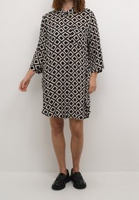 Sort og beige mønstret skjortekjole med diamantformer, knapper foran, lange ærmer og enkelt brystlomme; båret med sorte loafers.