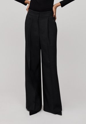 Wide-leg black trousers with a high waist, featuring pleats, a smooth texture, and precise tailoring for a structured fit.